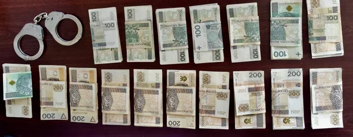 Rozłożone na ciemnym stole banknoty 100 i 200 złotych obok srebrnych kajdanek policyjnych.