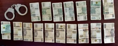 Rozłożone na ciemnym stole banknoty 100 i 200 złotych obok srebrnych kajdanek policyjnych.