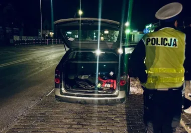 Policjant w kamizelce odblaskowej stoi przy samochodzie osobowym z otwartym bagażnikiem na brukowanej pobocznej drodze nocą.