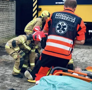Ratownik medyczny i strażacy udzielają pomocy poszkodowanemu leżącemu na ziemi przy karetkie.